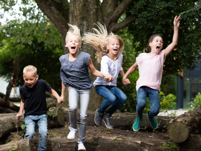 Vier Kinder springen fröhlich auf einem Baumstamm, umgeben von Bäumen.