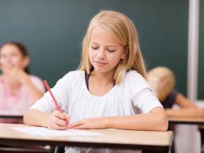 Ein Mädchen sitzt im Klassenzimmer und schreibt mit einem roten Bleistift auf einem Blatt Papier.