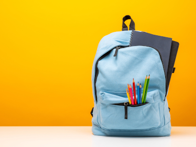 Ein blauer Rucksack mit bunten Buntstiften und einem Notizbuch vor einem gelben Hintergrund.