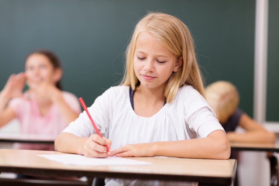 Ein Mädchen schreibt konzentriert in einem Klassenzimmer. Im Hintergrund sind weitere Kinder zu sehen.
